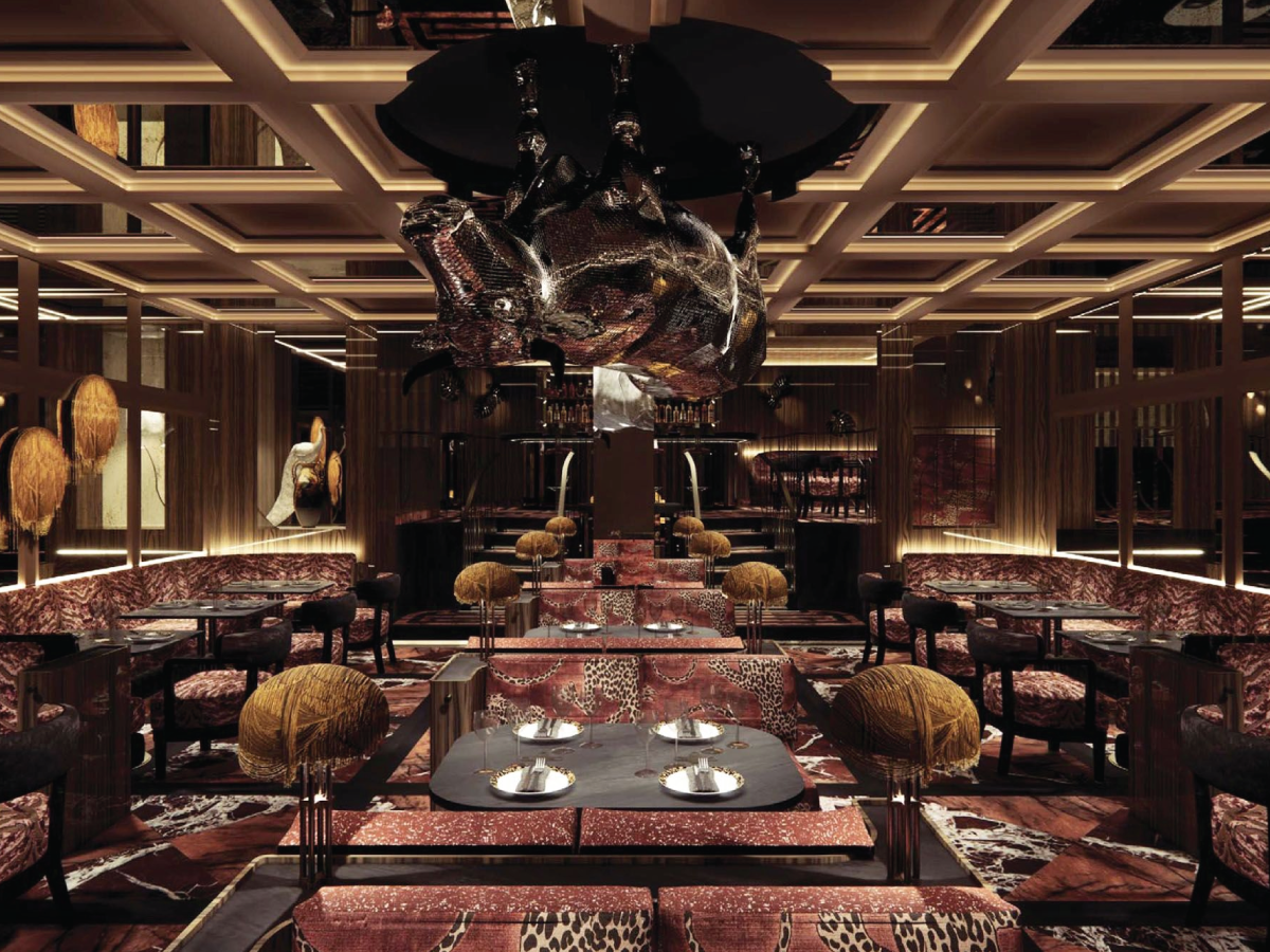 An ornate, luxurious restaurant interior with patterned red and brown upholstered seating, warm ambient lighting, geometric ceiling details, and a large reflective chandelier hanging from the center.