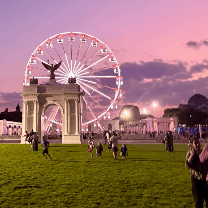A scenic view of a large illuminated Ferris wheel at dusk behind a white triumph arch, with people walking and playing on a grassy lawn in the foreground under a pink and purple sky.
