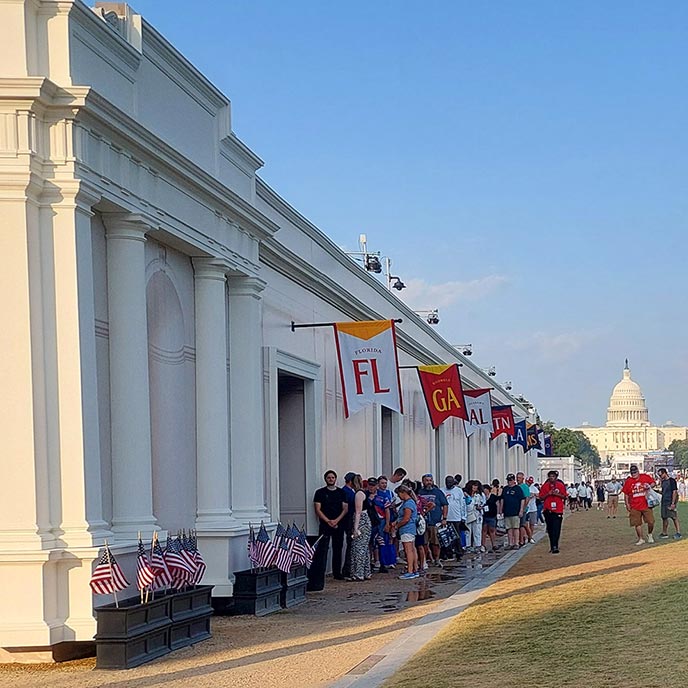 A long line of people stands outside a white building with columns under a blue sky, with the U.S.