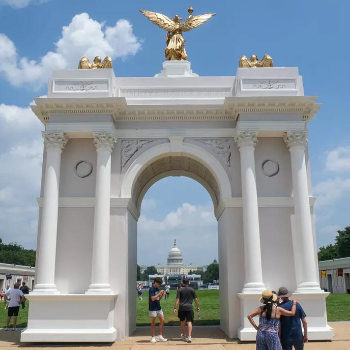 A large white triumphal arch with gold winged statues on top stands outdoors under a blue sky with clouds. People are walking through and around the arch, and a domed government building is visible in the distance.