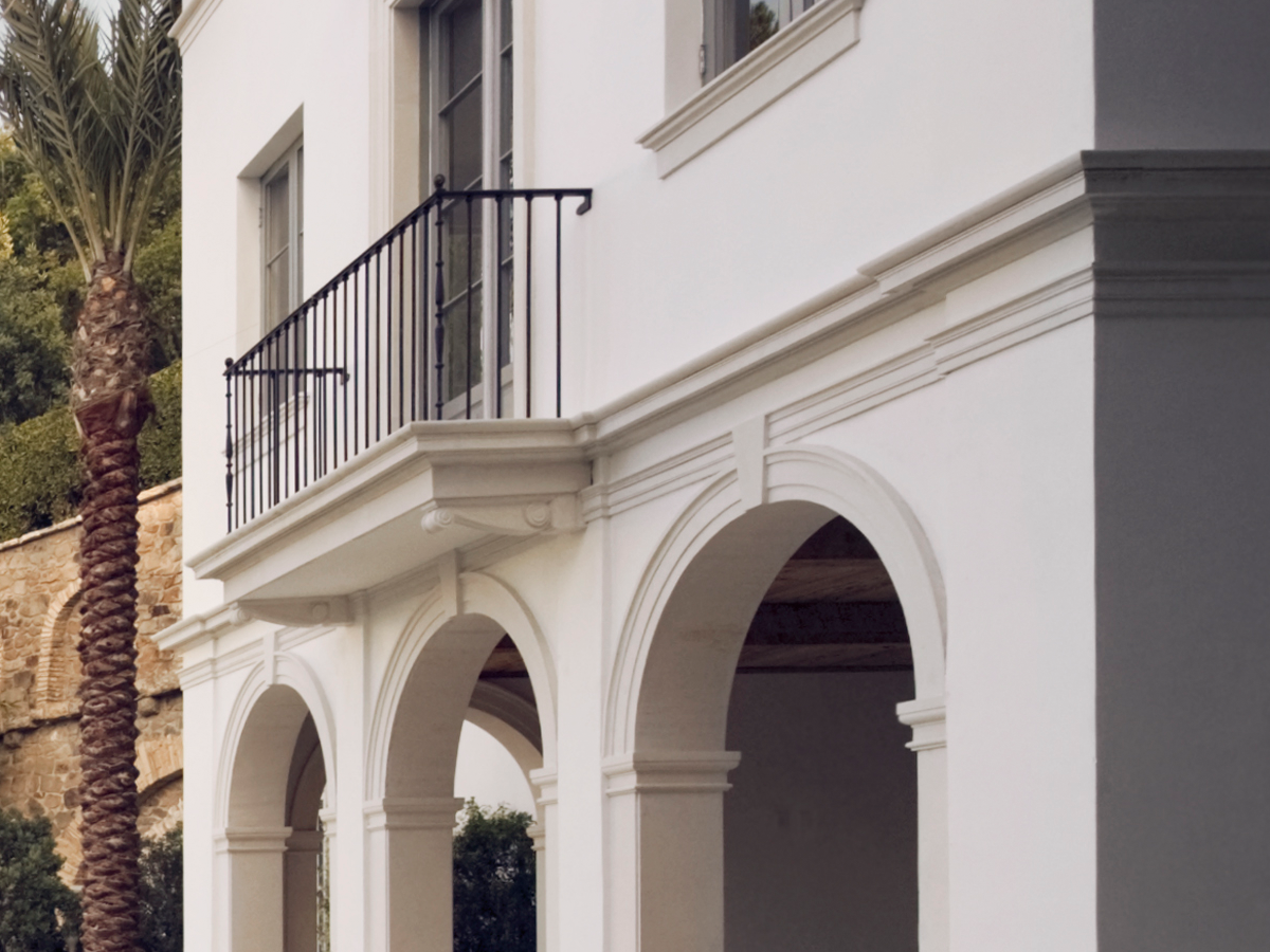 White stucco building facade with arched colonnade, black metal balcony railing, tall windows, and a palm tree at the left edge.