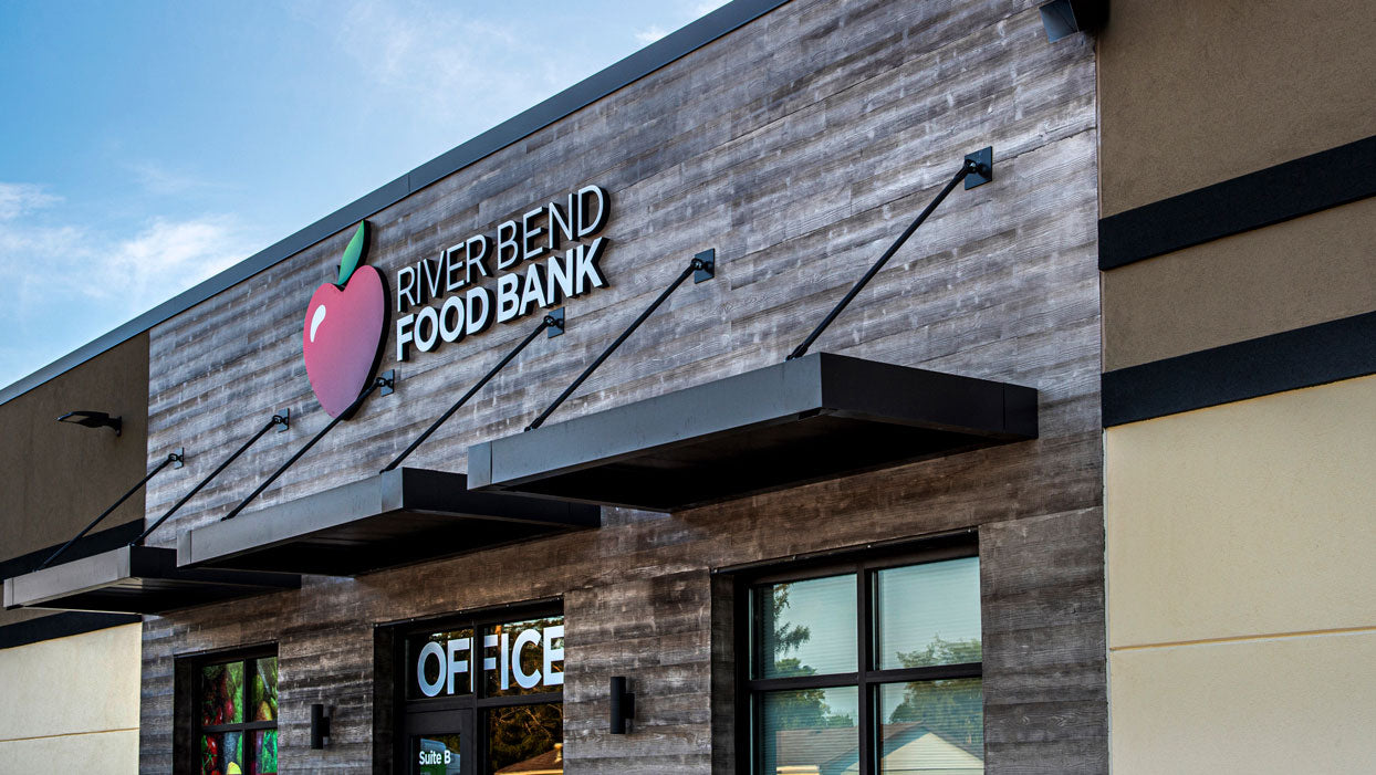 Commercial building storefront for River Bend Food Bank featuring weathered grey wood-grain composite wall panels and modern black metal awnings.